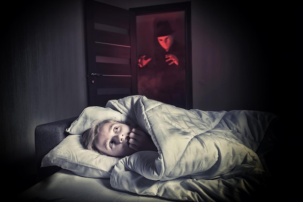 Nightmare Scared boy lying in the bed while the masked stranger standing in a doorway