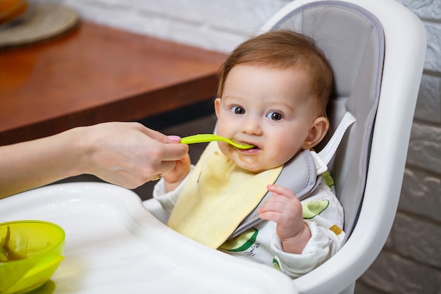 9개월 된 웃는 아기가 하이체어의 흰색 테이블에 앉아 그릇에서 숟가락으로 먹습니다. 엄마는 숟가락으로 아기를 먹입니다. 흐리게 배경입니다. 어린이를 위한 건강식. 어린이 음식.