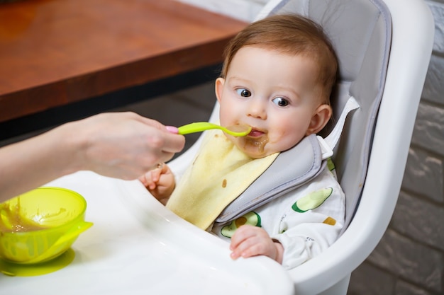 A nine month old smiling baby sits at a white table in a highchair and eats with a spoon from a bowl