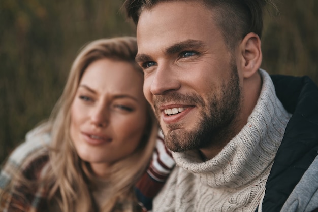 No cares.  Beautiful young couple looking away and smiling while sitting on the field outdoors