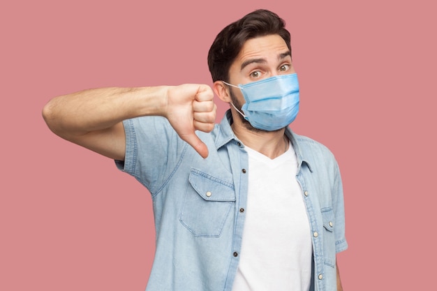 No, non mi piace. ritratto di giovane insoddisfatto con maschera medica chirurgica in camicia blu in piedi, pollice in giù e guardando la fotocamera. girato in studio al coperto, isolato su sfondo rosa.