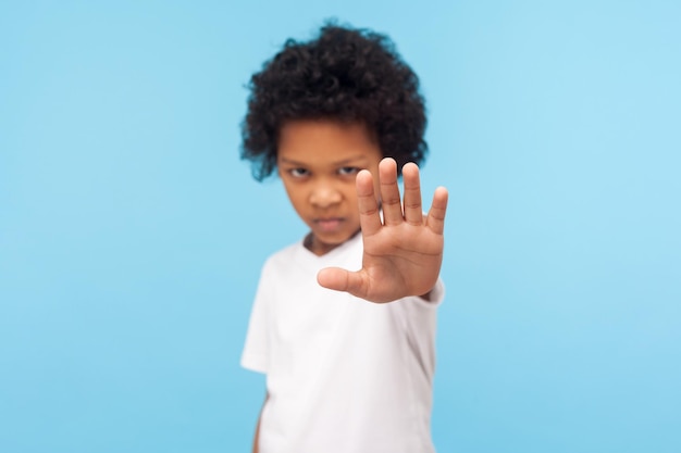 Photo no, forbidden. portrait of serious angry little boy with curly hair showing stop gesture, warning to go and looking aggressive, rejection denial concept. indoor studio shot isolated on blue background