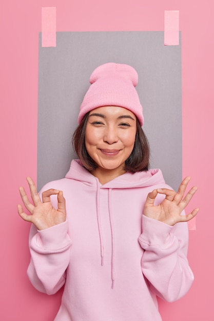 No problem. Pleased sincere Asian woman assures everything under control approves something good being assertive in her choice guarantees quality wears hat and casual sweatshirt poses indoor
