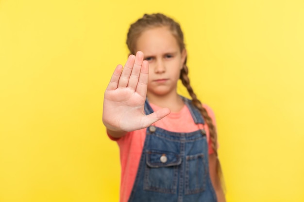 No, rejection sign. Portrait of unhappy little girl in denim overalls showing stop gesture, warning to go, looking angry dissatisfied, denial concept. indoor studio shot isolated on yellow background