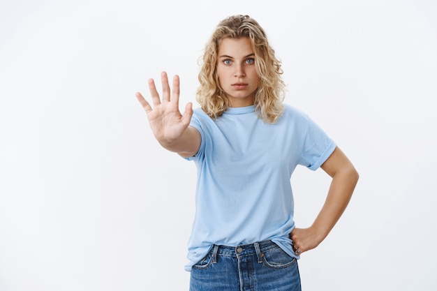 いいえ、やめてください。真面目そうな自信と勇気のある若い女性、短い巻き毛のブロンドの髪が手のひらをカメラに向かって引っ張ることは決してありません、禁止と十分なジェスチャー、白い壁の上に自信を持って