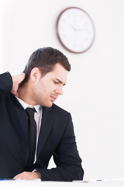 No time for break. Depressed young man in formalwear looking away and touching his neck while sitting at his working place with wall clock on background