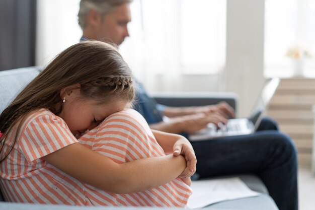 子供のための時間はありません。ラップトップで忙しい白髪のお父さん、家でオンラインで働いている、近くに座っている悲しい退屈な気分を害した娘。