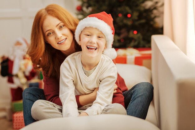 No time for worries. Loving mother hugging her excited son tightly while both sitting on a sofa and smiling at home.