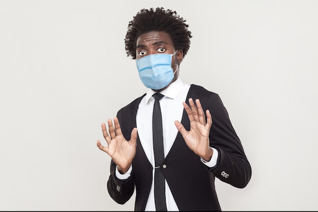 No, wait, stop please. Portrait of shocked or scared man wearing black suit with medical mask standing with blocking hands and looking with afraid face. indoor studio shot isolated on gray background