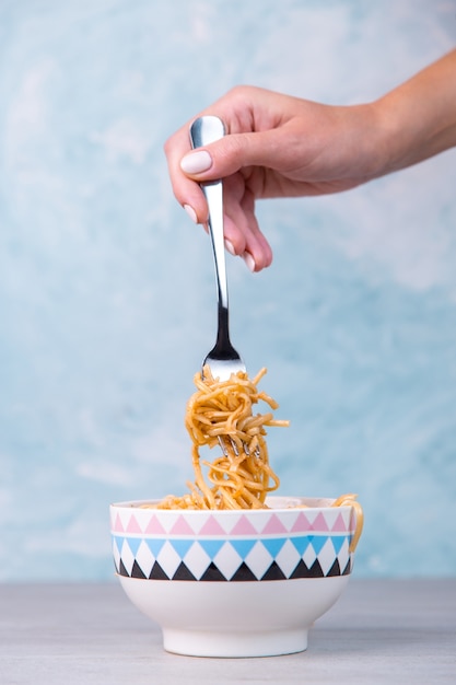 Noedels met saus in een gekleurde kom, hand houdt een vork van pasta opknoping, eetlust spaghetti op blauw.