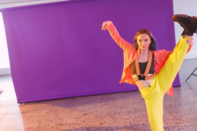 Nonbinary person performing high leg extension in studio with purple backdrop stand