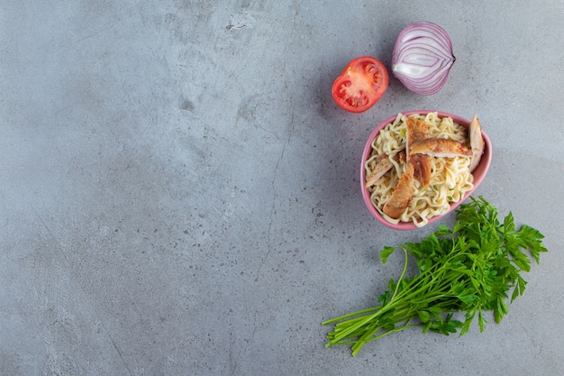 Noodle with meat in a bowl next to parsley bunch, tomatoes and onion, on the marble background. 