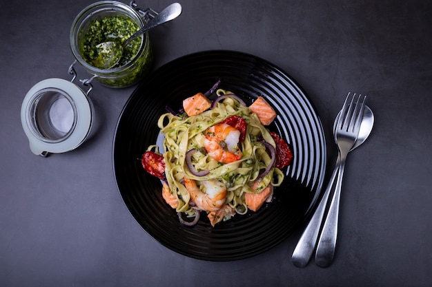 Noodles with seafood, sun-dried tomatoes, capers and red onions. Homemade pasta with shrimp, salmon (trout) and pesto sauce. Black background, black plate. Close-up.