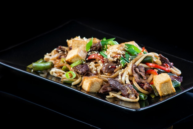 noodles with vegetables with pork, chicken, fish, scallop, beef, shrimp in a black plate on a black wall.