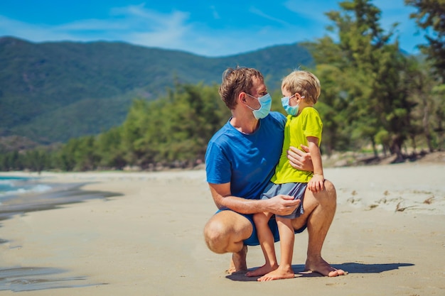 Normale realiteit tijdens COVID Vader die hurkt en zoon vasthoudt terwijl hij naar elkaar kijkt met een blauw gezichtsmasker, beschermt het coronavirus, zelfs wandelend in de natuur zand zee strand in de buurt van bosparkzondag