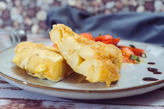 Normale weergave van een bord met twee porties Spaanse aardappelomelet en gesneden tomaten met olijf- en sherryazijn op een houten tafel