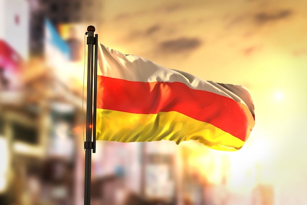 North Ossetia-Alania Flag Against City Blurred Background At Sunrise Backlight