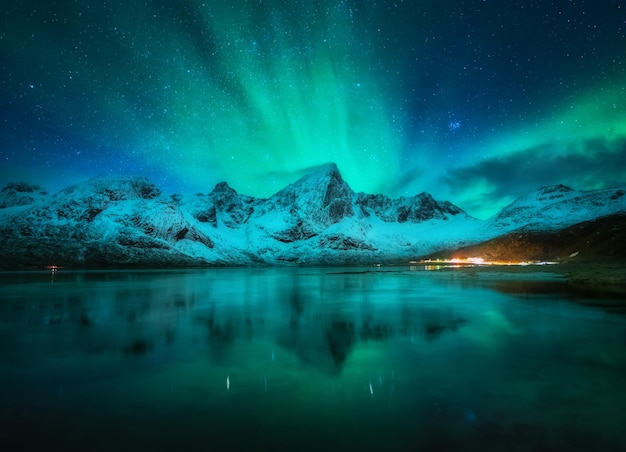 雪の山の上のオーロラ、ノルウェーのロフォーテン諸島の夜に水に映る凍った海のオーロラと雪に覆われた岩、極光の星空とフィヨルドのある冬の風景