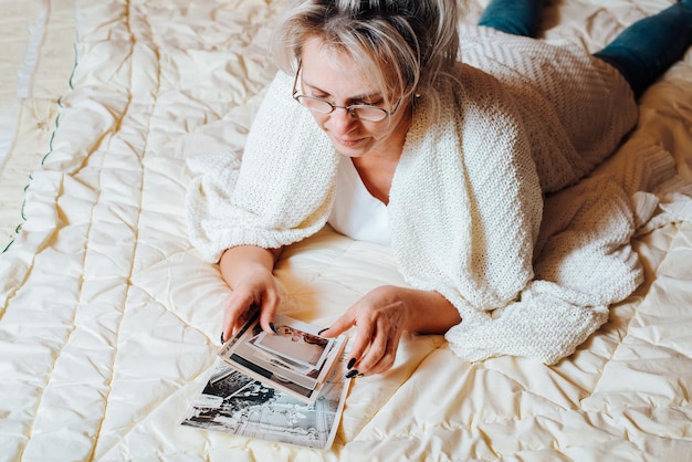 Nostalgia and memories concept. Middle-aged caucasian woman looking at old family album photos while relaxing on bed in bedroom, top view. Adult woman and vintage pictures of childhood and youth.