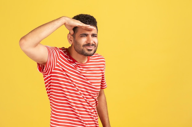 Nosy attentive man with beard in red striped t-shirt looking far away holding hand near forehead, watching at distance, searching for something. Indoor studio shot isolated on yellow background