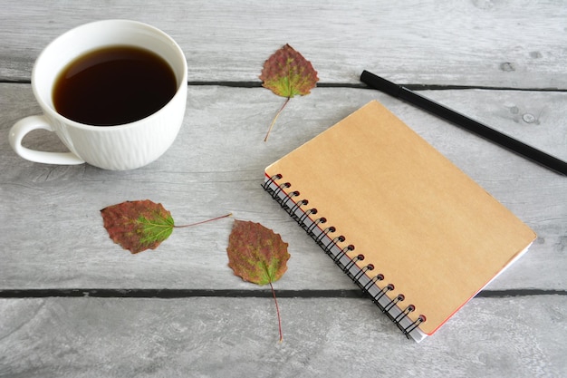 Taccuino e penna nera con tazza di caffè bianca e foglie autunnali secche su fondo di legno grigio