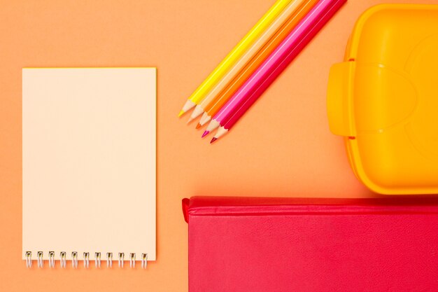 ノートブック、本、色鉛筆、ピンクの背景にランチボックス。コピースペースのある上面図。学校のコンセプトに戻ります。学用品。パステルカラー