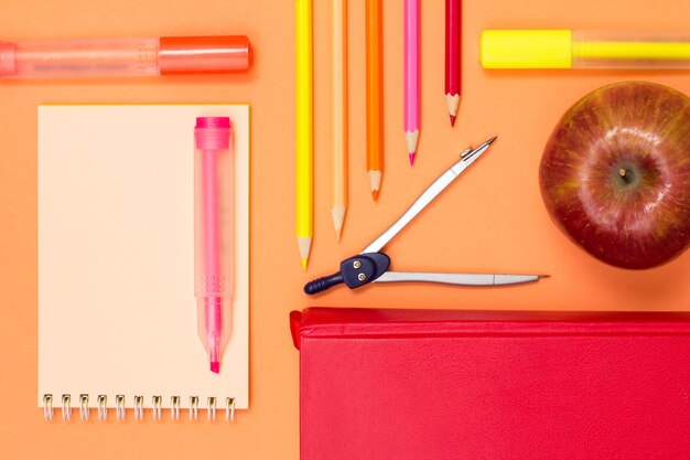 Photo notebook, book, compass, color pencils and apple on pink background.