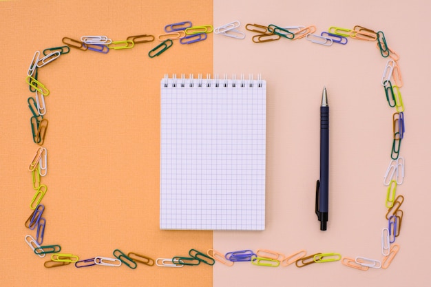 Notebook in cage with a pen and pencil in a frame of colored paper clips on a double background - beige and orange. concept back to school