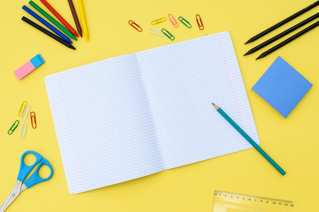 黄色の背景に鉛筆、消しゴム、定規、ペーパークリップ、その他の事務用品が入った檻の中のノート。学校に戻るコンセプト。テキストの場所。