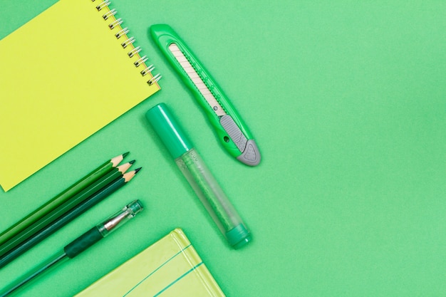 Notebook, kleurpotloden, pen, boek, viltstift en papiermes op groene achtergrond. Bovenaanzicht met kopie ruimte. Terug naar schoolconcept. Schoolspullen
