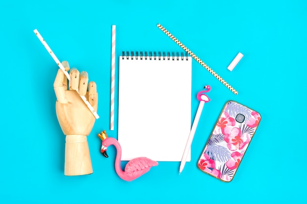 notebook, pen, flamingo figure, smartphone, wooden hand hold drinking paper straws on blue background