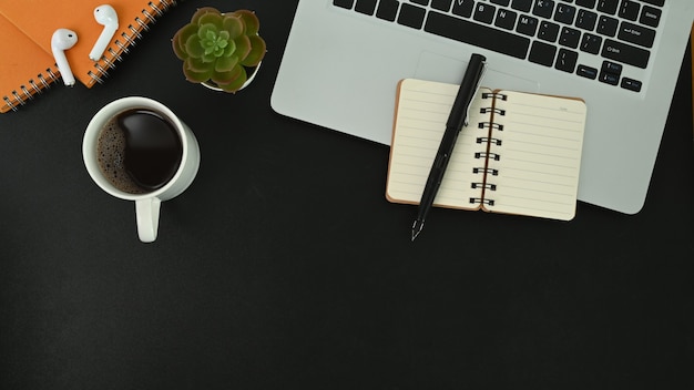 Notebook and pen over laptop computer, coffee cup, cactus an earphone on black leather.