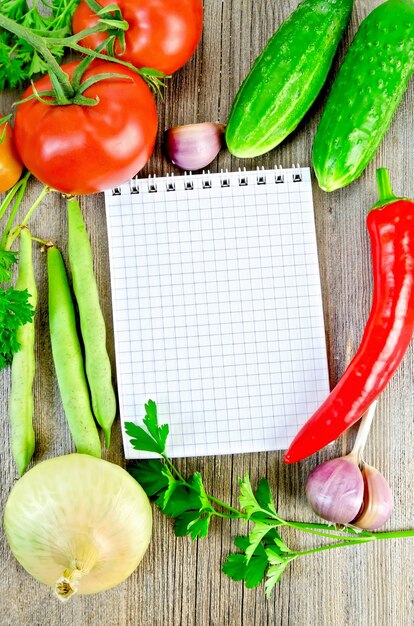 Notebook, red tomatoes, red hot pepper, parsley, garlic, onion, cucumber, green bean pods on an old wooden board