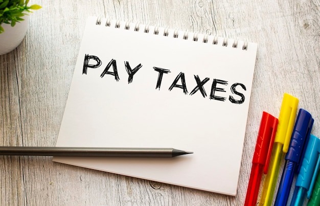 白いシートにPAYTAXESというテキストが書かれた春のノートは、色付きのペンが付いた木製のテーブルの上にあります。ビジネスコンセプト。