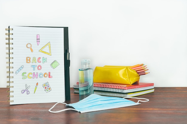 Notebook with back to school drawing. Study material, mask and gel on wooden table. Copy space