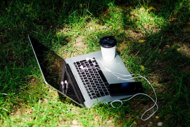 Notebook with headset of mobile phone and coffee to go on green grass summer vibes Relax university students life Online shopping agile business Online study working place outdoor Free music