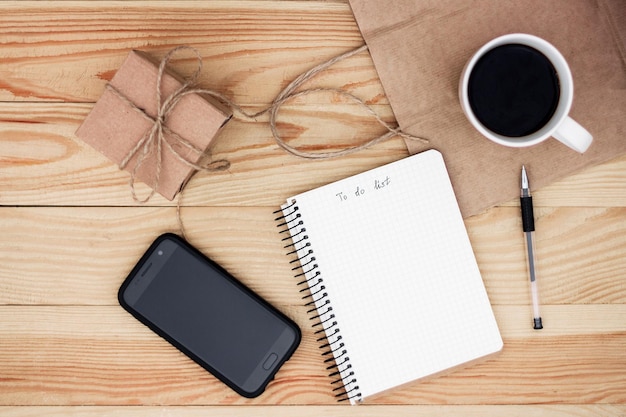 A notebook with an inscription To do list laying on wooden table. It is surrounded by phone, black pen, cup of coffee, present and a paper bag.