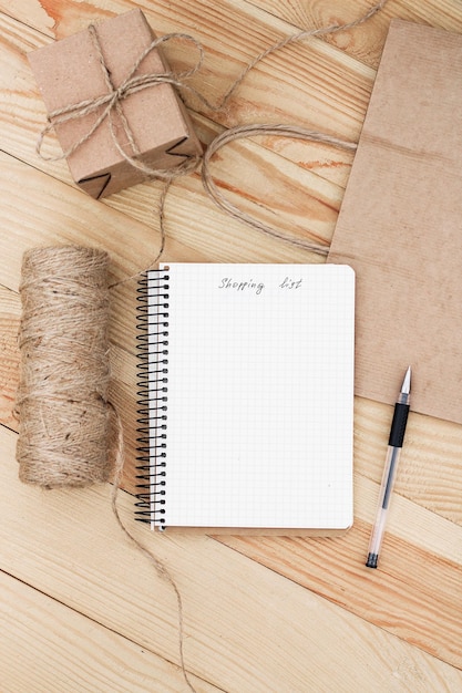 A notebook with an inscription Shopping list  laying on wooden table. It is surrounded by   present, black pen, rope twine and a paper bag.