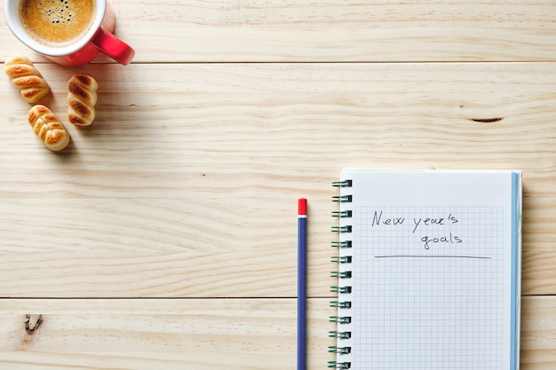 新年の目標が木製の背景に書かれ、その横に鉛筆があり、左上隅に赤いコーヒーが入ったノートブック
