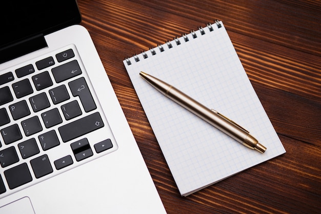 A notebook with a pen lies next to a laptop on a wooden table.