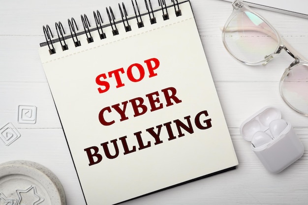 Foto taccuino con frase stop cyber bullying occhiali e auricolari su tavolo di legno bianco piatto