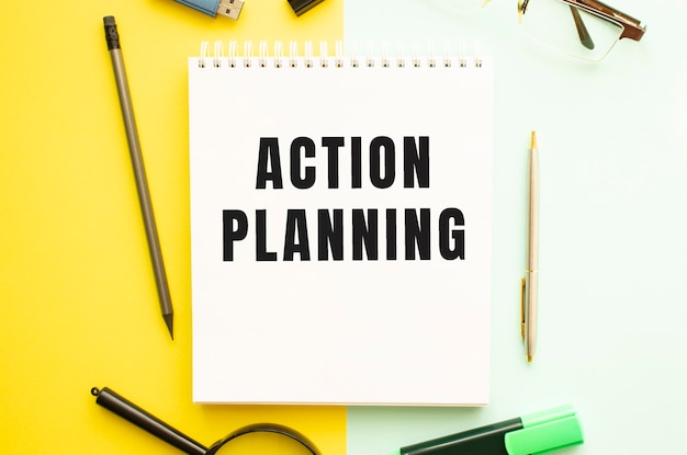 事務用品を備えたオフィステーブルに「ACTION PLANNING」というテキストが書かれたノート。黄色の背景のコンセプト。