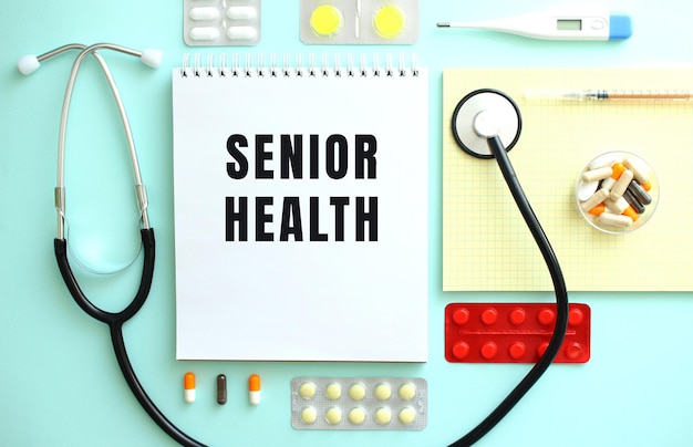 A notebook with the text SENIOR HEALTH is neatly folded among the pills a stethoscope and a yellow notebook
