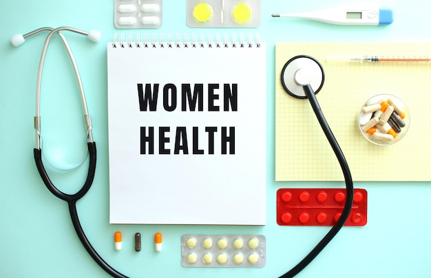 A notebook with the text WOMEN HEALTH is neatly folded among the pills a stethoscope and a yellow notebook