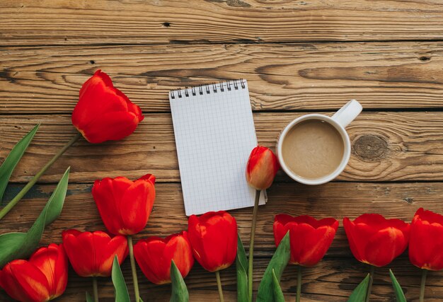 Notebook with white pages, red tulips and cup of coffee on the wooden background.