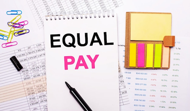 EQUAL PAY라는 단어가있는 노트북, 마커, 색종이 클립 및 밝은 메모 용지가 그래프 표면에 놓여 있습니다.