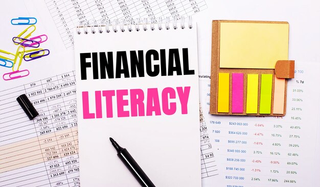 FINANCIAL LITERACY라는 단어가있는 노트북, 마커, 색종이 클립 및 밝은 메모 용지가 그래프 배경에 놓여 있습니다.