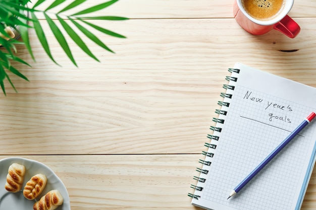 Notebook with the words new year's goals written on a natural wooden background with a blue and red pencil on top, some green leaves next to it and a cup of coffee in the corner