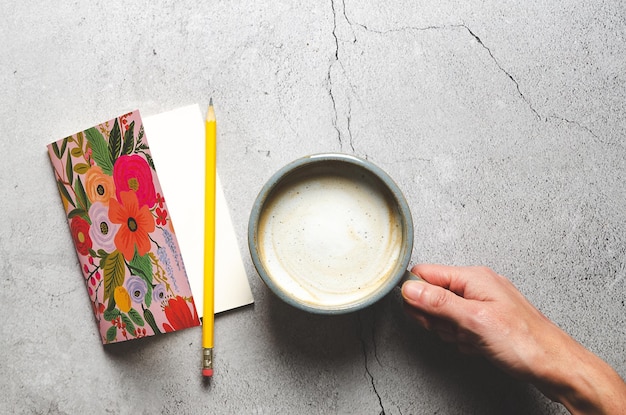 A notebook with a yellow pencil and a right hand holding a cup of latte on a grey stoned background.