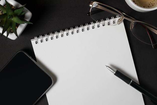 Notebooks with empty white sheets black pen mobile phone glasses and cup of coffee on a dark background copy space Business or education concept